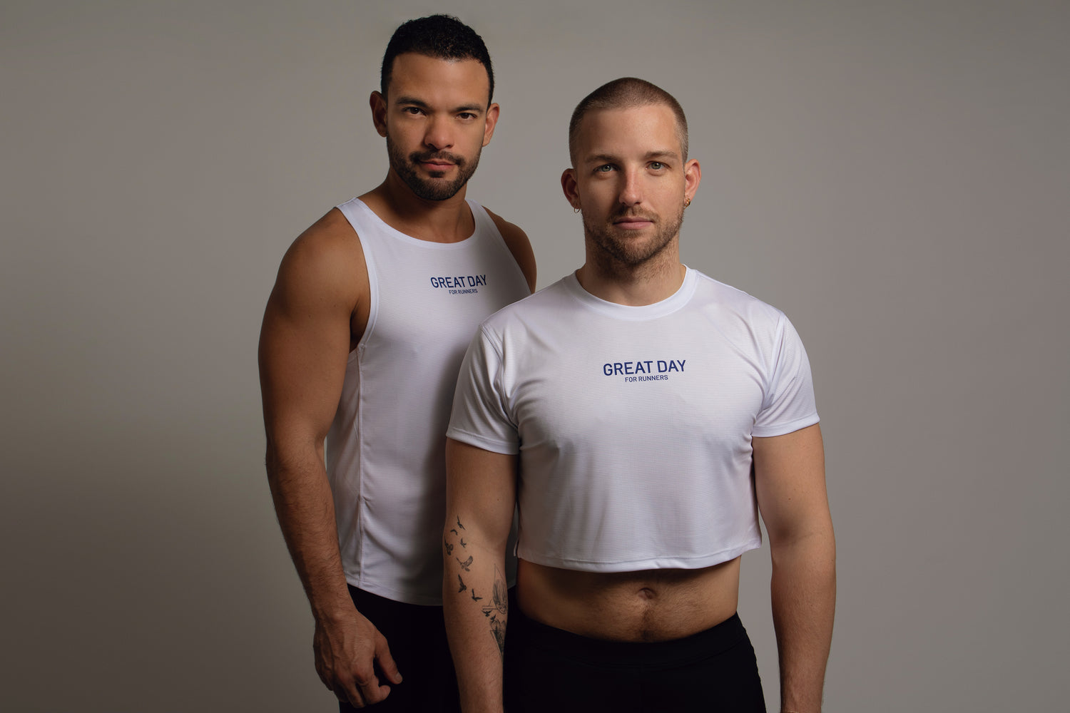 Two men in white crop tops standing against a grey background, both with text "great day" on his shirt, looking confidently at the camera.